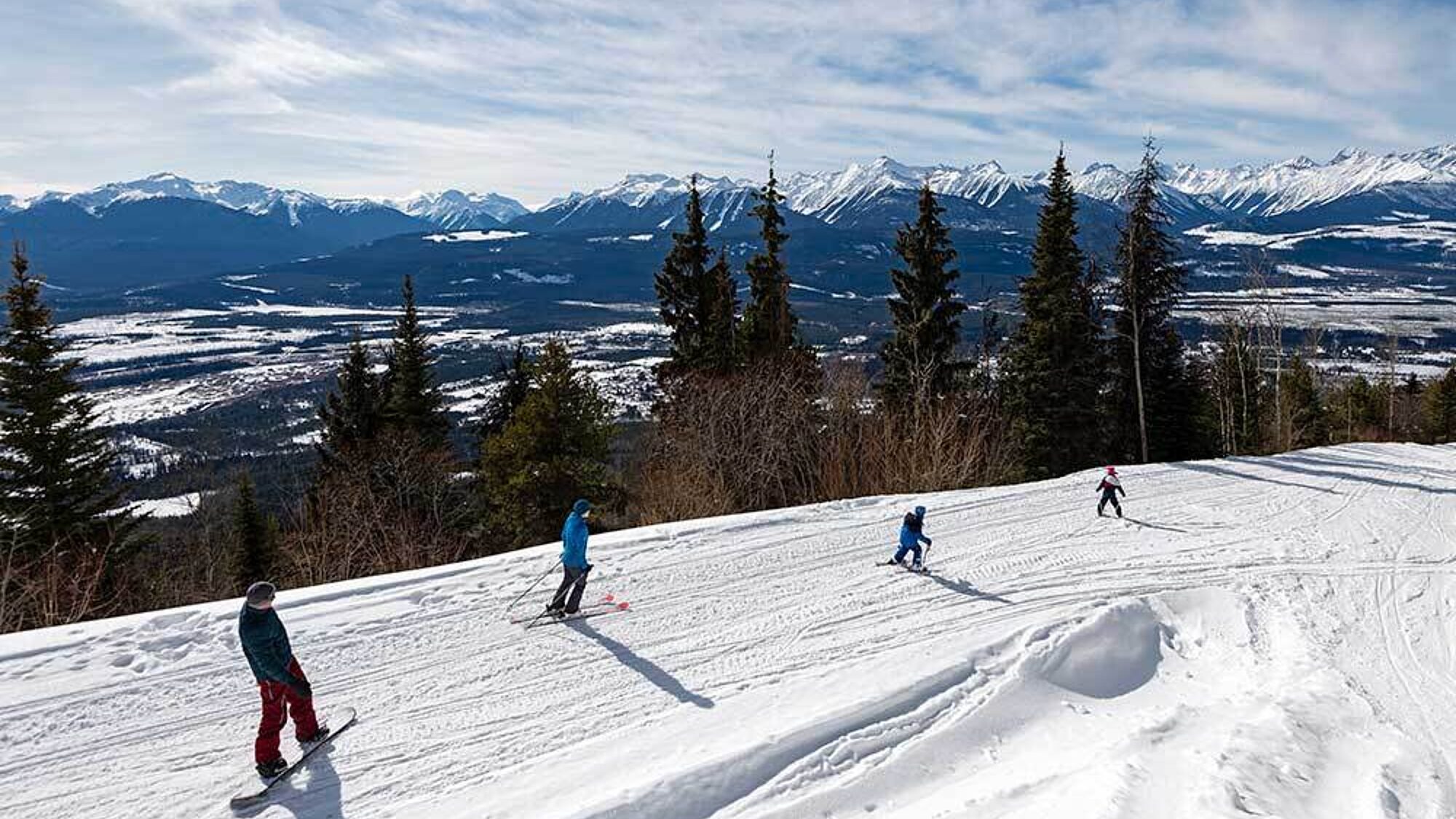 People snowmobiling downhill on a wide snow-covered slope with mountain views